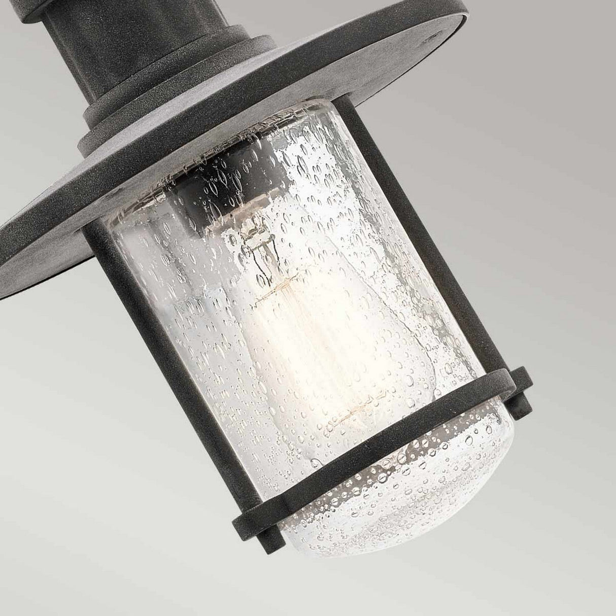 Close-up of the Riverwood Outdoor 1 Light Chain Pendant showcasing a weathered zinc frame and clear, textured glass cover. Inside, a bulb glows against a neutral gray background, exuding rustic outdoor lighting charm.