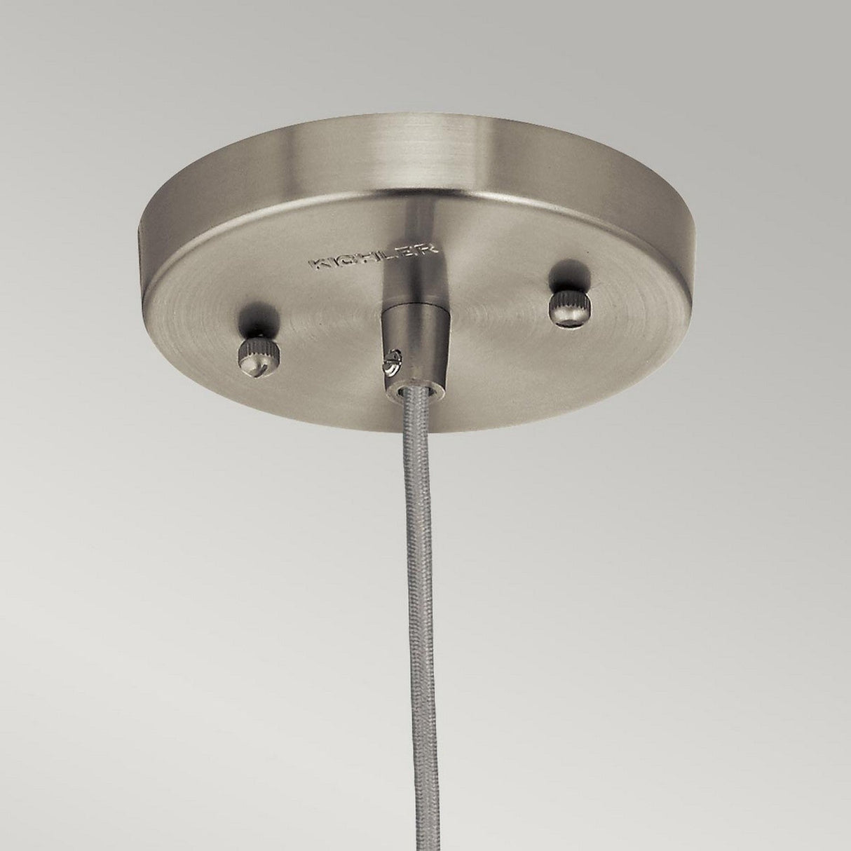 The Riviera-1 1 Light Pendant - Brushed Nickel features a brushed nickel ceiling canopy with a smooth finish. Its cylindrical fixture and vintage-inspired design, complete with a single cord that descends gracefully, embody minimalist elegance enhanced by two discreet screws.