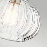 A close-up of the Riviera-1 1 Light Pendant - Brushed Nickel, showcasing its vintage-inspired design with an exposed incandescent bulb. It emits a warm glow, highlighted by a fluted clear glass and complemented by the brushed nickel finish against the soft gray background.