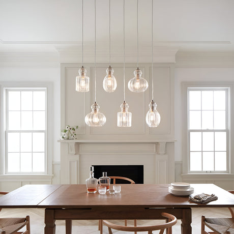 A dining room featuring a wooden table is adorned with glasses and plates, while overhead, six Riviera-1 1 Light Pendants in Brushed Nickel elegantly hang with glass shades of various shapes. In the background, large windows and a white fireplace mantel enhance the room's charm.