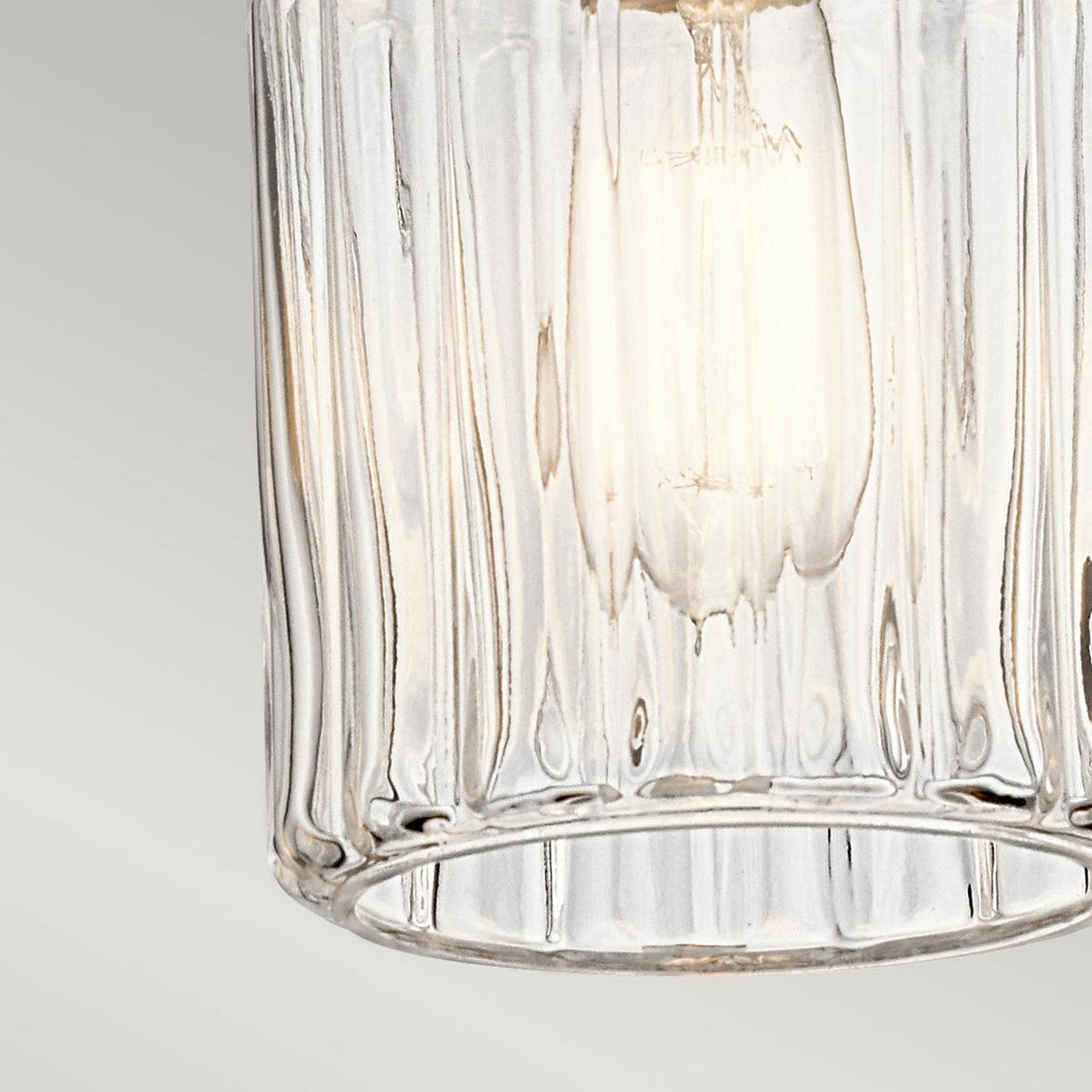 Close-up of the Riviera-2 1 Light Pendant in Brushed Nickel, featuring a clear, textured glass shade. The bulb inside emits a warm glow, creating a cozy ambiance over the kitchen island. The background is a plain, gradient grey.