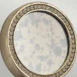 The Rosalie 1 Light Wall Light in Sterling Gold showcases a round mirror with a metal frame accented by intricate floral patterns. The mirror glass, reflecting an antique mercury finish, displays charming signs of age with cloudy spots.