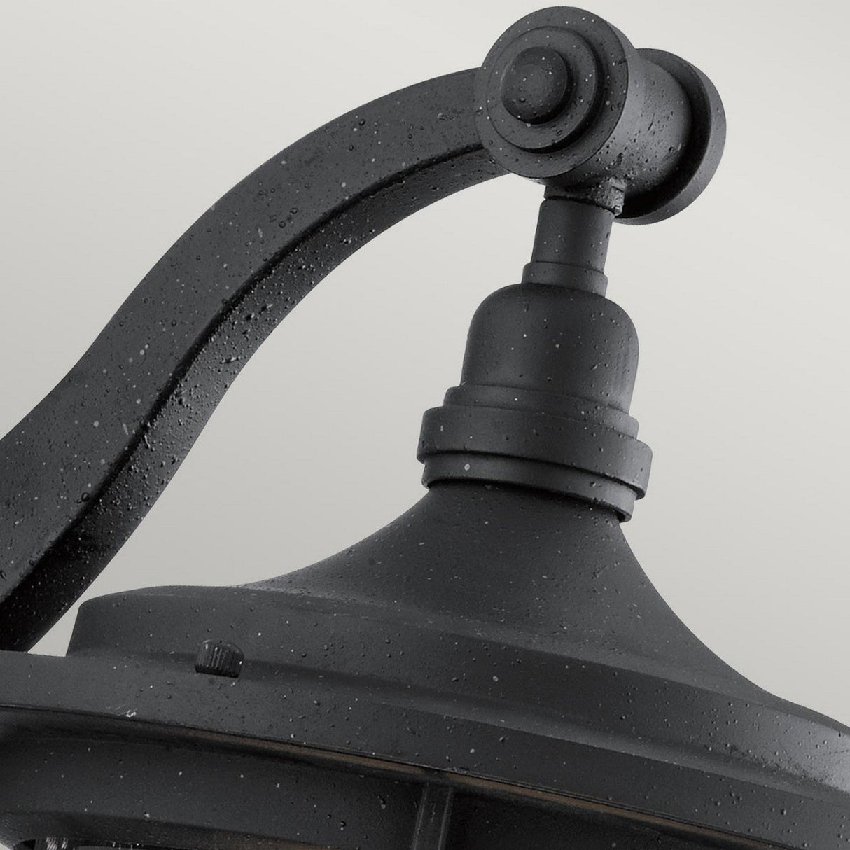Close-up of the Royal Marine Outdoor 1 Light Medium Wall Lantern in black, featuring a curved arm and round top that enhance its timeless vintage appeal. The surface glistens with droplets from recent rain, set against a plain light gray background that highlights this charming piece of outdoor lighting.