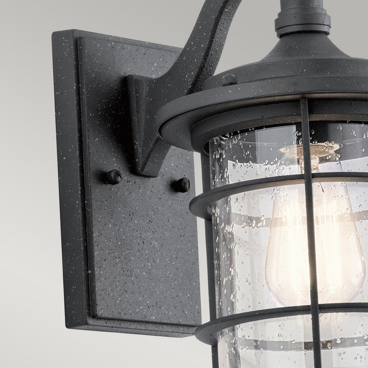 The Royal Marine Outdoor 1 Light Small Wall Lantern in black features a close-up view of its seeded glass, which displays a clear raindrop pattern. Inside, the bulb casts a warm light. The fixture is installed on a rectangular backplate with a simple, modern design that subtly enhances its nautical appearance.