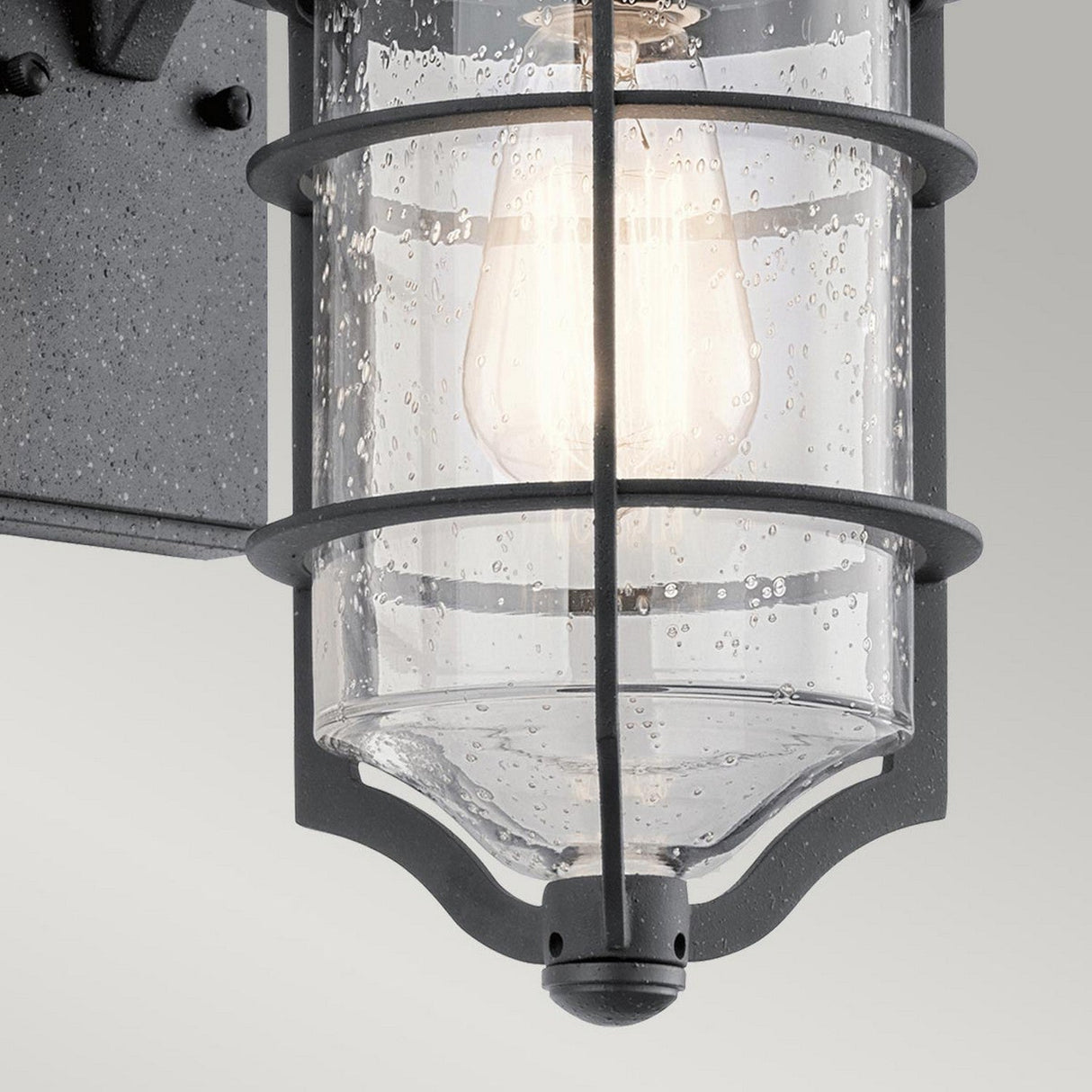 A close-up view of the Royal Marine Outdoor 1 Light Small Wall Lantern in black, featuring a metal frame and clear seeded glass. The illuminated bulb casts enchanting, rain-like shadows. This wall lantern adds a nautical touch to the textured wall it embellishes.