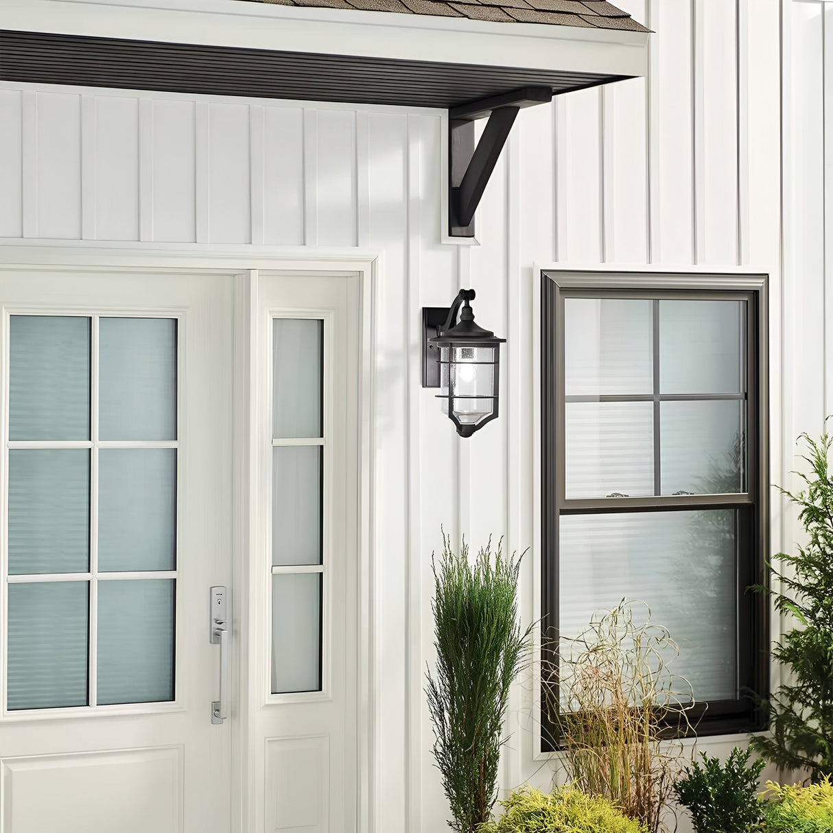 The exterior of the house showcases a modern white door with glass panels and is complemented by the Royal Marine Outdoor 1 Light Medium Wall Lantern in black, which adds a touch of vintage charm. A window with sleek black trim completes the look. Surrounding the entrance are green plants and shrubs, providing a striking contrast to the immaculately white walls.