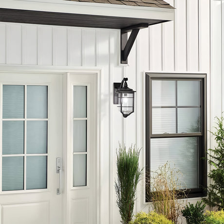 The exterior of the house showcases a modern white door with glass panels and is complemented by the Royal Marine Outdoor 1 Light Medium Wall Lantern in black, which adds a touch of vintage charm. A window with sleek black trim completes the look. Surrounding the entrance are green plants and shrubs, providing a striking contrast to the immaculately white walls.