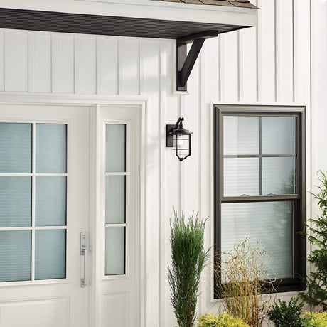 A minimalist house exterior showcasing a white door with vertical seeded glass panels is complemented by a window with a black frame and grid design. Above the small garden filled with various plants, enhancing the nautical aesthetic, is mounted the Royal Marine Outdoor 1 Light Small Wall Lantern in black.