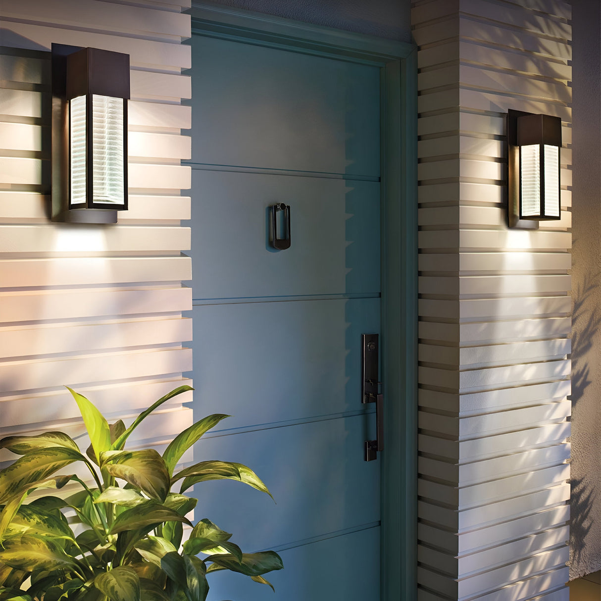 A teal front door with a silver handle is set against a light-colored, horizontally paneled wall. Two Sorel Outdoor 1 Light Wall Lanterns in bronze illuminate the doorway, and a potted plant with green leaves is positioned on the left.