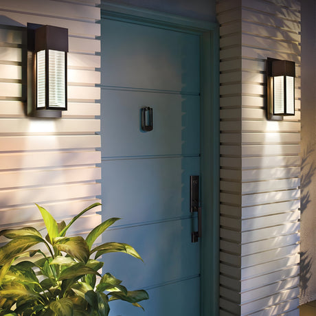 A teal front door with a silver handle is set against a light-colored, horizontally paneled wall. Two Sorel Outdoor 1 Light Wall Lanterns in bronze illuminate the doorway, and a potted plant with green leaves is positioned on the left.
