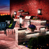 An outdoor patio with brick walls at sunset showcases a modern grill, potted plants, and South Hope Outdoor 1 Light Medium Wall Lanterns in rubbed bronze. Nearby is a table with navy chairs, set against the sky’s gradient of orange and purple hues for a warm ambiance.