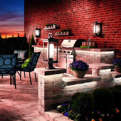 An outdoor patio with brick walls at sunset showcases a modern grill, potted plants, and South Hope Outdoor 1 Light Medium Wall Lanterns in rubbed bronze. Nearby is a table with navy chairs, set against the sky’s gradient of orange and purple hues for a warm ambiance.