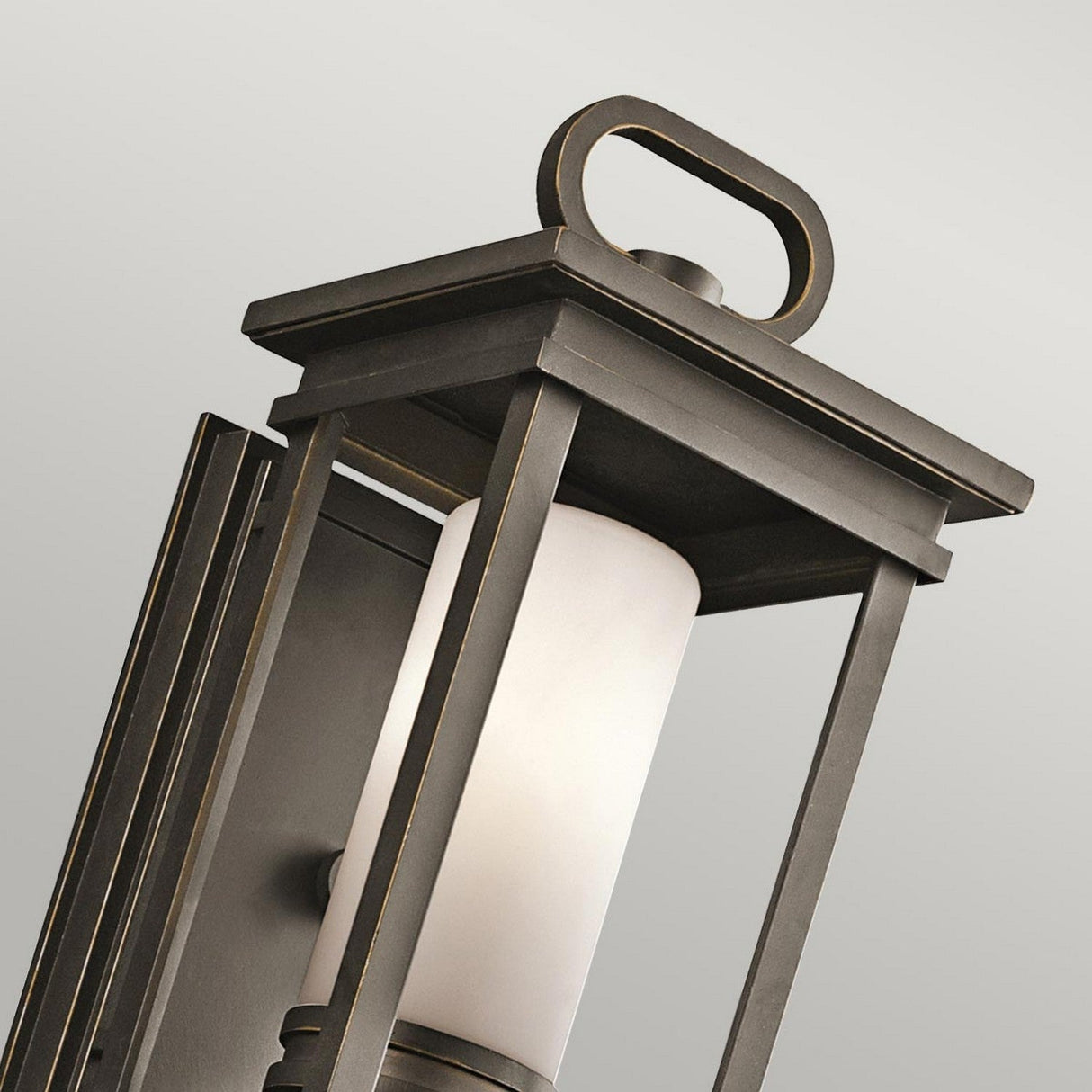 Close-up of the South Hope Outdoor 1 Light Small Wall Lantern in a rubbed bronze finish. This modern lantern showcases a rectangular frame with a handle-like detail on top, encasing an opal glass cylindrical light diffuser, set against a neutral background.
