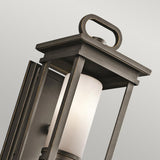 Close-up of the South Hope Outdoor 1 Light Small Wall Lantern in a rubbed bronze finish. This modern lantern showcases a rectangular frame with a handle-like detail on top, encasing an opal glass cylindrical light diffuser, set against a neutral background.
