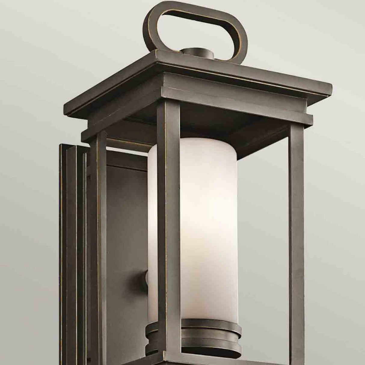 The South Hope Outdoor 1 Light Small Wall Lantern in rubbed bronze showcases a black metal rectangular frame with a loop handle and elegantly houses an opal glass cylindrical light fixture within. When installed on a pale gray background, it provides both sophistication and illumination.