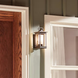 The South Hope Outdoor 1 Light Small Wall Lantern in rubbed bronze is installed on the exterior wall of a house, next to a wooden door and a window adorned with white curtains. The lantern's opal glass shade projects warm light, enhancing the cozy ambiance.