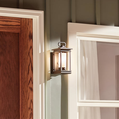 The South Hope Outdoor 1 Light Small Wall Lantern in rubbed bronze is installed on the exterior wall of a house, next to a wooden door and a window adorned with white curtains. The lantern's opal glass shade projects warm light, enhancing the cozy ambiance.