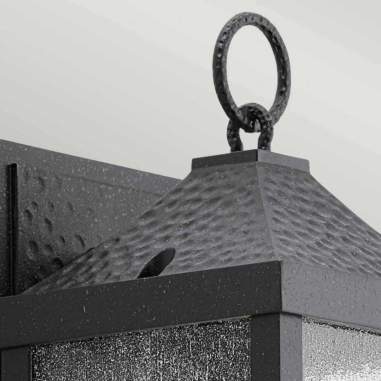 Close-up of the Springfield Outdoor 1 Light Small Wall Lantern, showcasing a textured, distressed black finish and a loop ring at the top. This square design includes clear glass panels accented with water droplet patterns. IP44 rated for outdoor use, it is mounted on a metal wall bracket.