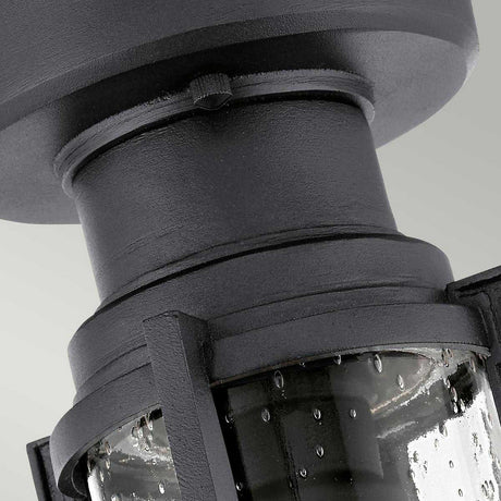Close-up of the Suri Outdoor 1 Light Porch/Pedestal Lantern in black, showcasing its textured lantern design with a cylindrical glass cover adorned with water droplet patterns. This fixture, radiating nautical charm, is mounted against a light gray background.