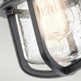 Close-up of the Suri Outdoor 1 Light Porch/Pedestal Lantern in black with a textured metal frame and clear glass shade. The warm glow of the bulb enhances its rustic vintage appeal with nautical charm.