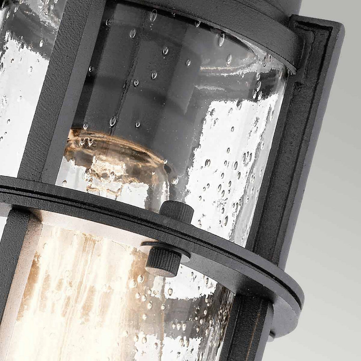 A close-up view of the Suri Outdoor 1 Light Porch/Pedestal Lantern in black showcases its cylindrical glass speckled with water droplets, encasing a glowing bulb. The textured black frame contrasts elegantly with the clear, textured glass, offering a sleek, modern design with a touch of nautical charm.
