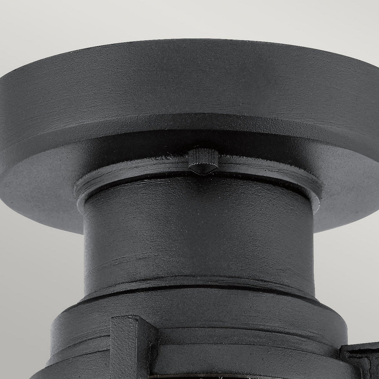 A close-up view of the Suri Outdoor 1 Light Porch/Pedestal Lantern in black, showcasing its textured cylindrical metal structure with a wide, flat top. The smooth surface and clean lines evoke an industrial or architectural element with nautical charm, set against a neutral gray background.