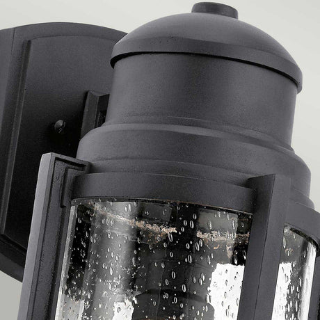 The Suri Outdoor 1 Light Wall Lantern in black showcases a close-up of its cylindrical glass shade with a bubbled texture. This lighting fixture is mounted on a rectangular base, combining modern and sleek design elements against a pale background.