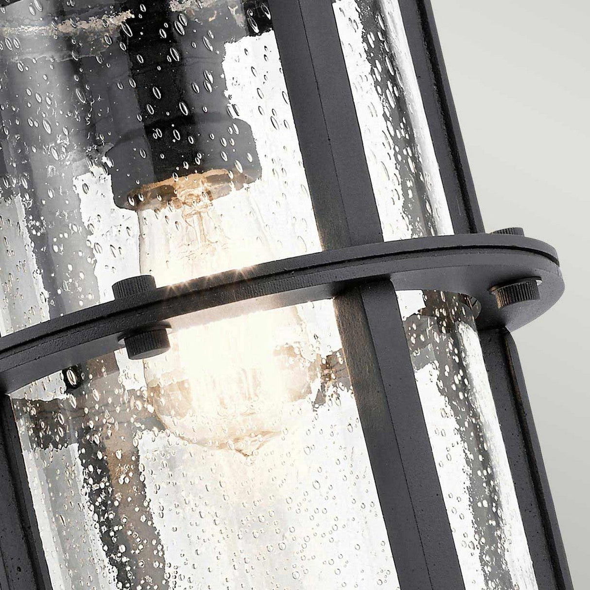 A close-up showcases the Suri Outdoor 1 Light Wall Lantern in black, featuring a clear glass cylindrical cover adorned with droplets. Inside, a glowing bulb emits a warm light. The dark-finished metal frame, inspired by nautical design, adds charm and offers UV protection for lasting appeal.