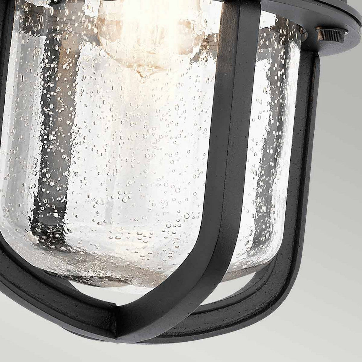 Close-up of the Suri Outdoor 1 Light Wall Lantern in black, featuring a nautical-inspired design with a textured glass shade encased in a metal frame. The bubbled texture enhances its vintage aesthetic, reminiscent of classic ship lighting. The lantern softly illuminates the space.