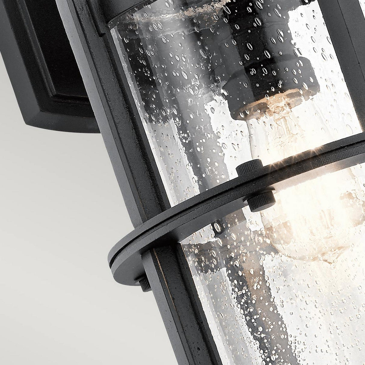 A close-up of the Suri Outdoor 1 Light Wall Lantern in Black showcases its cylindrical design with a glass cover speckled with raindrops. This nautical-inspired piece features a glowing light bulb that emits a warm glow through the wet glass. Mounted against a light gray background, it adds charm to your outdoor lighting setup.