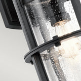 A close-up of the Suri Outdoor 1 Light Wall Lantern in Black showcases its cylindrical design with a glass cover speckled with raindrops. This nautical-inspired piece features a glowing light bulb that emits a warm glow through the wet glass. Mounted against a light gray background, it adds charm to your outdoor lighting setup.