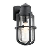The Suri Outdoor 1 Light Wall Lantern in black features a nautical-inspired design with a clear glass shade and metal details. Protective bars encase the cylindrical glass, revealing the visible bulb inside. This fixture is mounted on a rectangular wall plate, making it perfect for outdoor lighting.