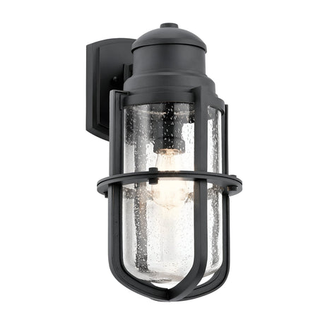 The Suri Outdoor 1 Light Wall Lantern in black features a nautical-inspired design with a clear glass shade and metal details. Protective bars encase the cylindrical glass, revealing the visible bulb inside. This fixture is mounted on a rectangular wall plate, making it perfect for outdoor lighting.