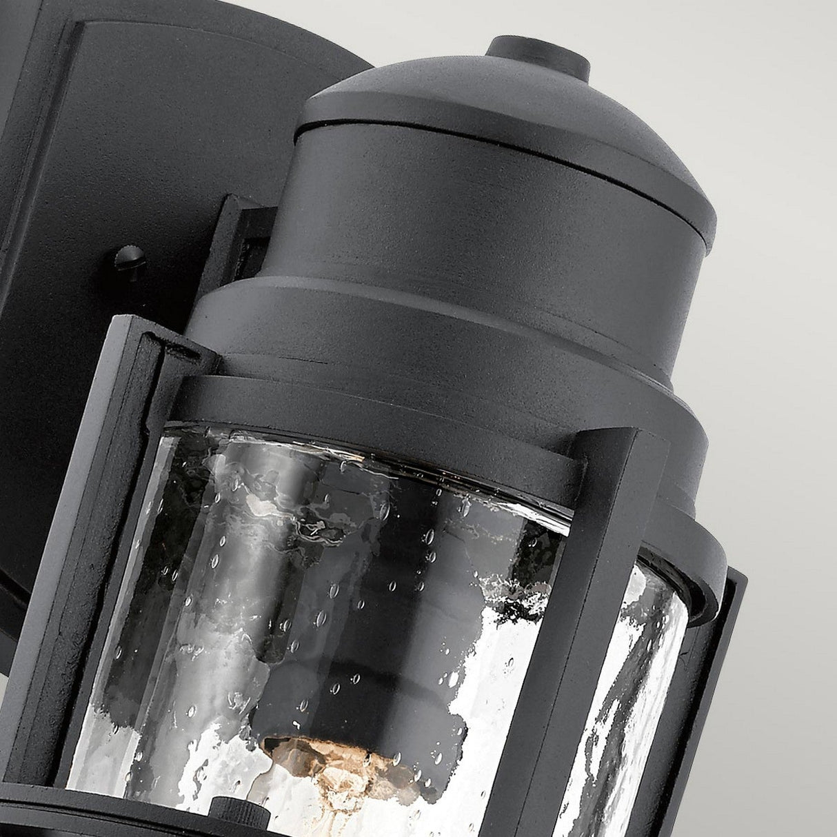 A close-up of the Suri Outdoor 1 Light Medium Wall Lantern - Black displays its clear, textured glass enclosure and sturdy metal frame. The light bulb inside is visible through the glass, emphasizing its design. Featuring coastal-inspired lighting aesthetics and UV protection, it stands out against the neutral background.