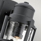 A close-up of the Suri Outdoor 1 Light Medium Wall Lantern - Black displays its clear, textured glass enclosure and sturdy metal frame. The light bulb inside is visible through the glass, emphasizing its design. Featuring coastal-inspired lighting aesthetics and UV protection, it stands out against the neutral background.