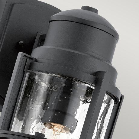 A close-up of the Suri Outdoor 1 Light Medium Wall Lantern - Black displays its clear, textured glass enclosure and sturdy metal frame. The light bulb inside is visible through the glass, emphasizing its design. Featuring coastal-inspired lighting aesthetics and UV protection, it stands out against the neutral background.