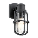 The Suri Outdoor 1 Light Small Wall Lantern - Black features a stylish design with a black wall-mounted frame and clear glass shade. Its cylindrical shape, metal cage, and exposed bulb evoke a modern industrial aesthetic with hints of coastal charm.