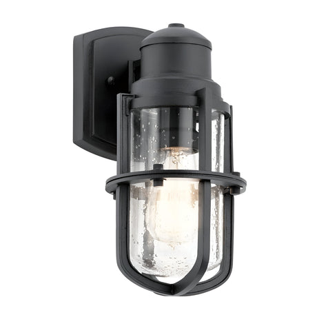The Suri Outdoor 1 Light Small Wall Lantern - Black features a stylish design with a black wall-mounted frame and clear glass shade. Its cylindrical shape, metal cage, and exposed bulb evoke a modern industrial aesthetic with hints of coastal charm.