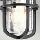 Close-up of the Suri Outdoor 1 Light Small Wall Lantern in black, exuding a vintage industrial style. It showcases a cylindrical glass cover encased in a metal cage, with its speckled texture gently revealing the bulb inside and casting a warm glow reminiscent of coastal design lanterns.