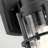 A close-up of the Suri Outdoor 1 Light Small Wall Lantern in black reveals raindrops on its clear glass housing. Its sleek and modern design brings a contemporary aesthetic that is ideal for enhancing any setting, be it an urban terrace or a coastal garden.