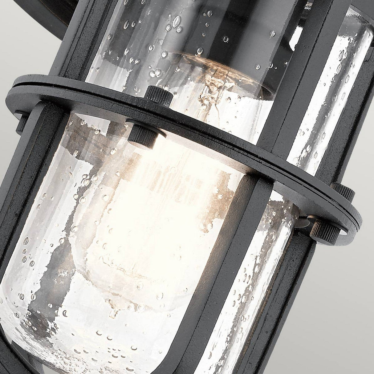A close-up view of the Suri Outdoor 1 Light Small Wall Lantern in black showcases its metal and glass construction. The textured bubble glass reveals a glowing bulb inside, emitting a warm light. This sophisticated fixture blends modern industrial style with coastal design charm, creating a captivating outdoor atmosphere.