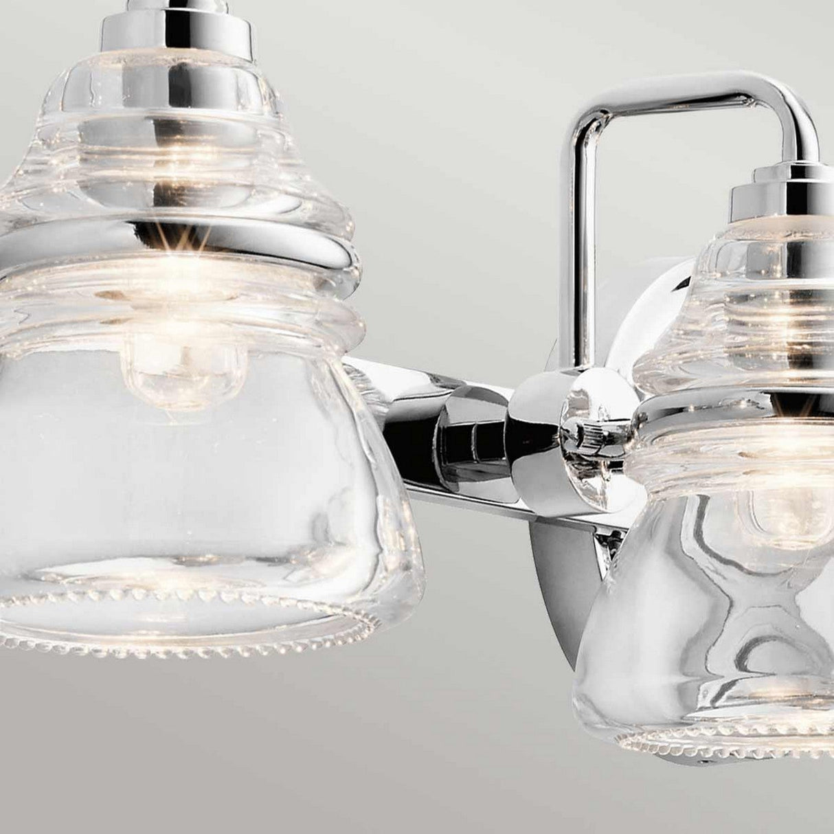 A close-up of the Talland 4 Light Wall Light in polished chrome showcases its modern design, featuring clear vintage glass shades and a reflective metal fixture against a plain background, making it perfect for bathroom lighting.