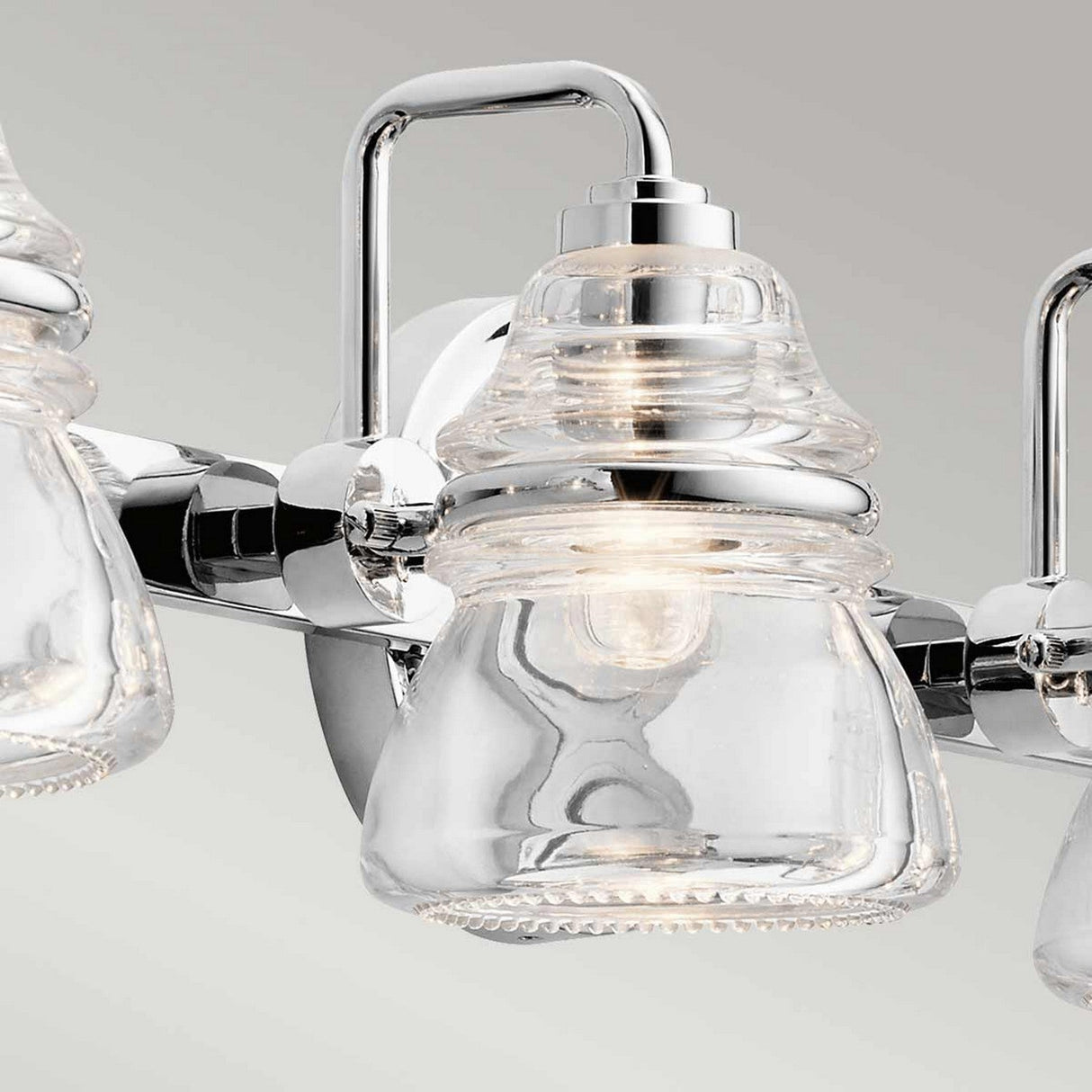 Introducing the Talland 4 Light Wall Light in polished chrome, featuring a close-up of its sleek design with a clear vintage glass shade. This fixture is elegantly mounted on a metallic bar with a smooth, reflective surface, seamlessly blending industrial and contemporary elements. Ideal for bathroom lighting, it brings an elegant touch to any space.