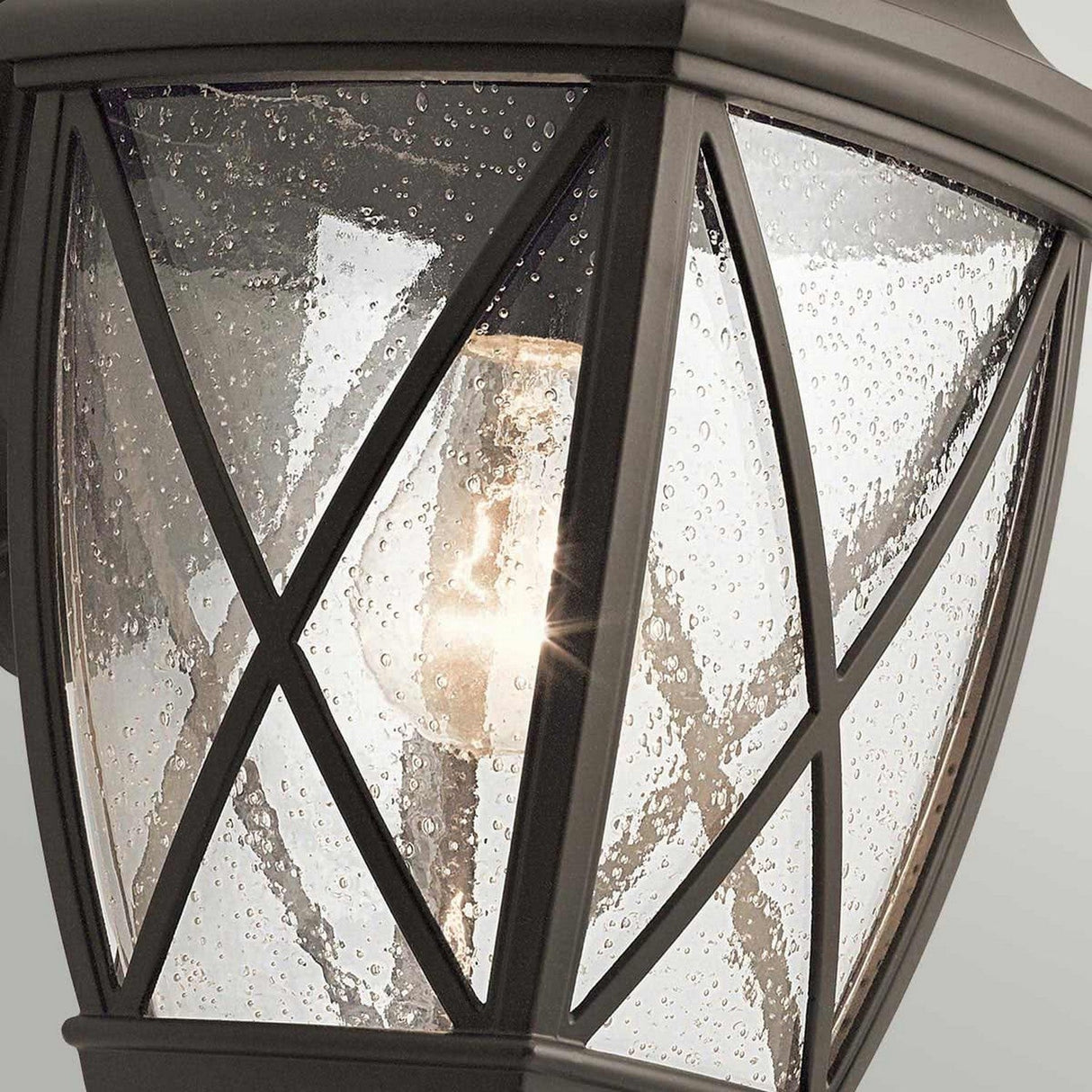 Close-up of the Tangier Outdoor 1 Light Medium Wall Lantern in Olde Bronze with raindrops on the glass. The light is illuminated, casting a warm glow. Its metal frame features a diagonal lattice design, adding a decorative touch and offering an IP44 rating for weather resistance.