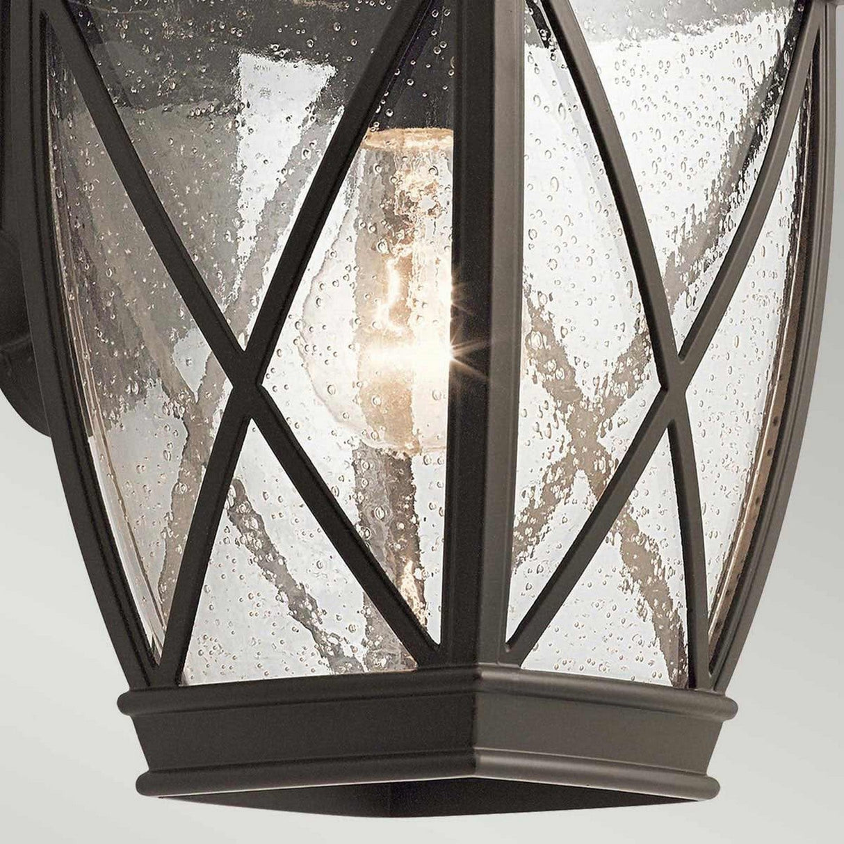 Close-up of the Tangier Outdoor 1 Light Medium Wall Lantern in Olde Bronze, featuring a black metal frame and clear glass panels textured with water droplets. Inside, a glowing bulb emits a warm light, and the lantern boasts an IP44 rating for weather-resistant durability.