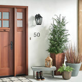 You'll find a cozy entryway featuring a wooden front door with glass panels, complemented by the Tangier Outdoor 1 Light Medium Wall Lantern in an Olde Bronze finish. A small bench holds boots and a decorative lantern, while potted plants and a rug complete the welcoming scene, all enhanced by the wall lantern's IP44 rating for durability.