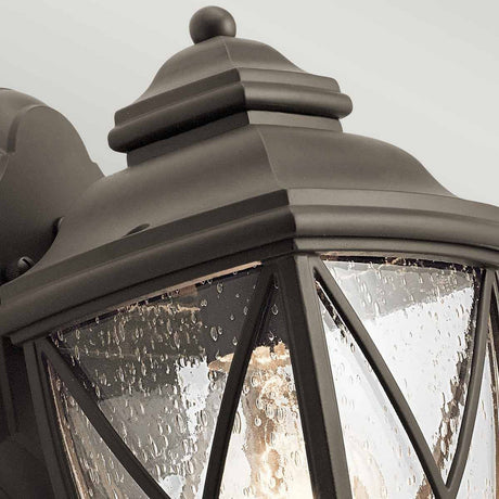 A close-up of the Tangier Outdoor 1 Light Small Wall Lantern in Olde Bronze showcases its vintage style. This small wall lantern is designed with textured glass panels featuring a water droplet pattern and crisscross metal details. Elegantly installed on a light gray wall, it enhances the charm of your outdoor lighting setup.