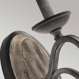 Close-up of the Taulbee 1 Light Wall Light, showcasing a rustic metal wall sconce with a weathered zinc finish and a circular wooden backplate. The design combines farmhouse decor with industrial elements, featuring a curved arm that supports an upward-facing candle holder. The background is a soft gray.