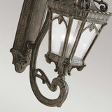 A close-up of the Tournai Outdoor 4 Light Grand Wall Lantern - Bronze highlights its intricate metalwork and classic design. The lantern boasts a dark bronze finish paired with seedy glass panels, elegantly affixed to a decorative wall mount.
