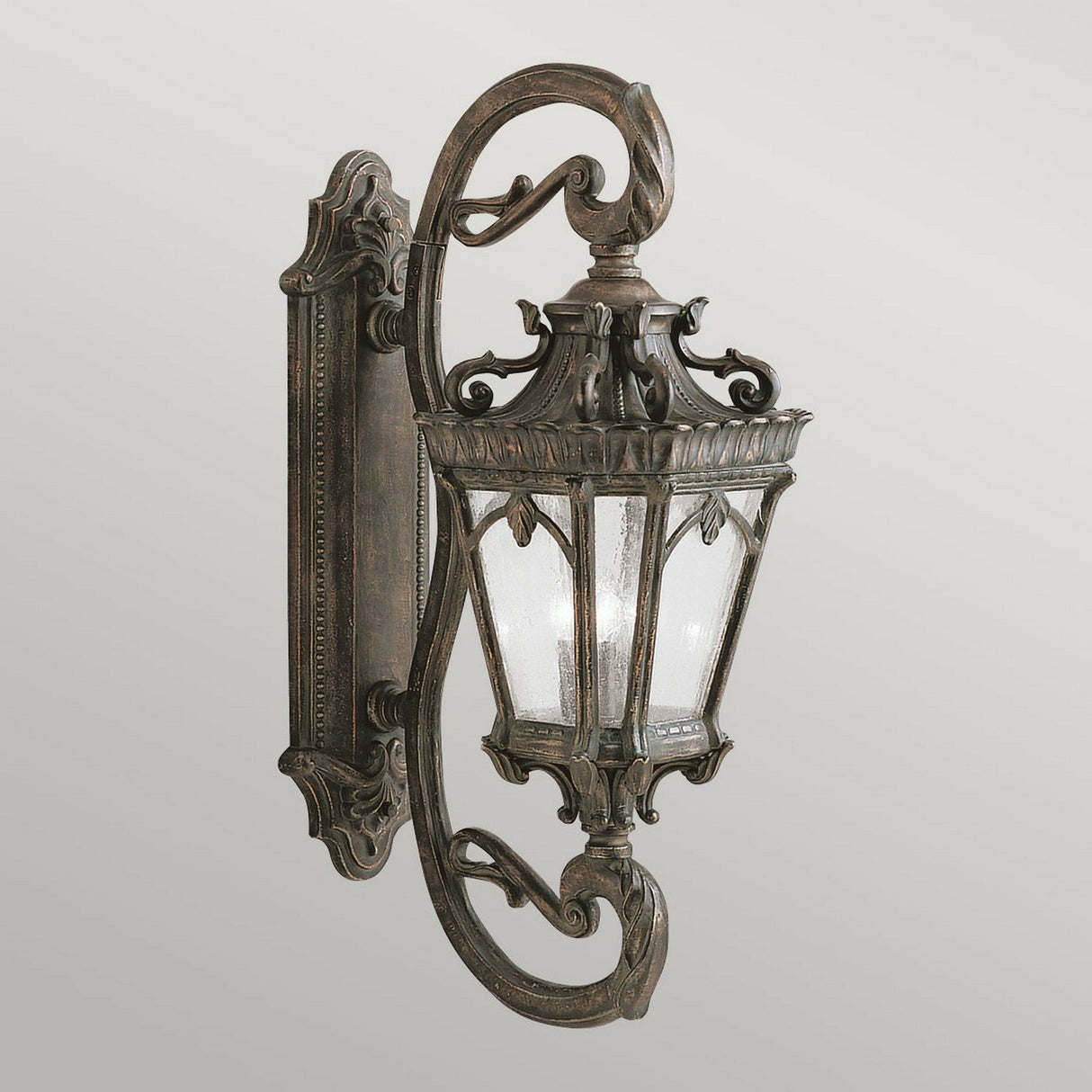 The Tournai Outdoor 4 Light Grand Wall Lantern - Bronze is a vintage-style outdoor fixture, showcasing intricate scrollwork and seedy glass panels within a glass-paneled enclosure. It is mounted on a decorative wall bracket and crafted from metal with an antique bronze finish.